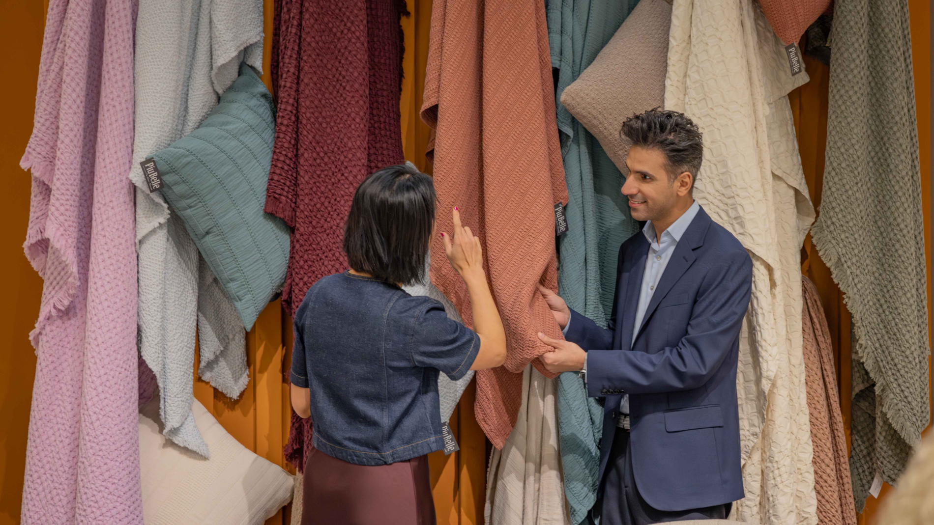 Two people examine and feel various colorful textiles hanging on a wall at an exhibition stand